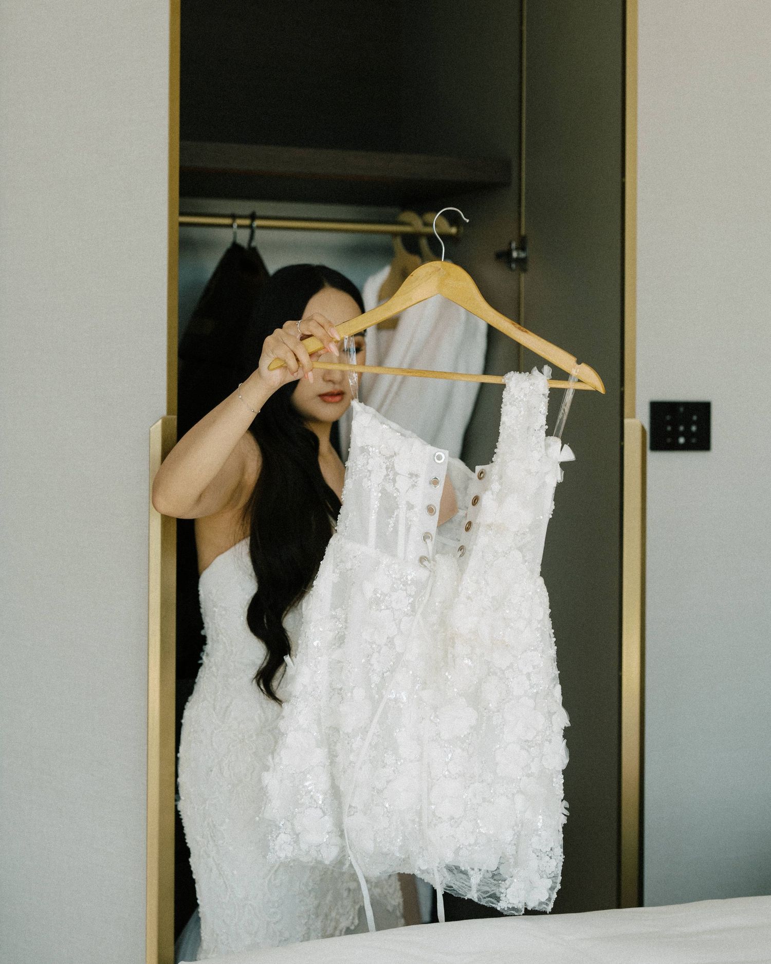 Someone wearing a black top holds up a white lace wedding dress on a hanger while looking at it excitedly.