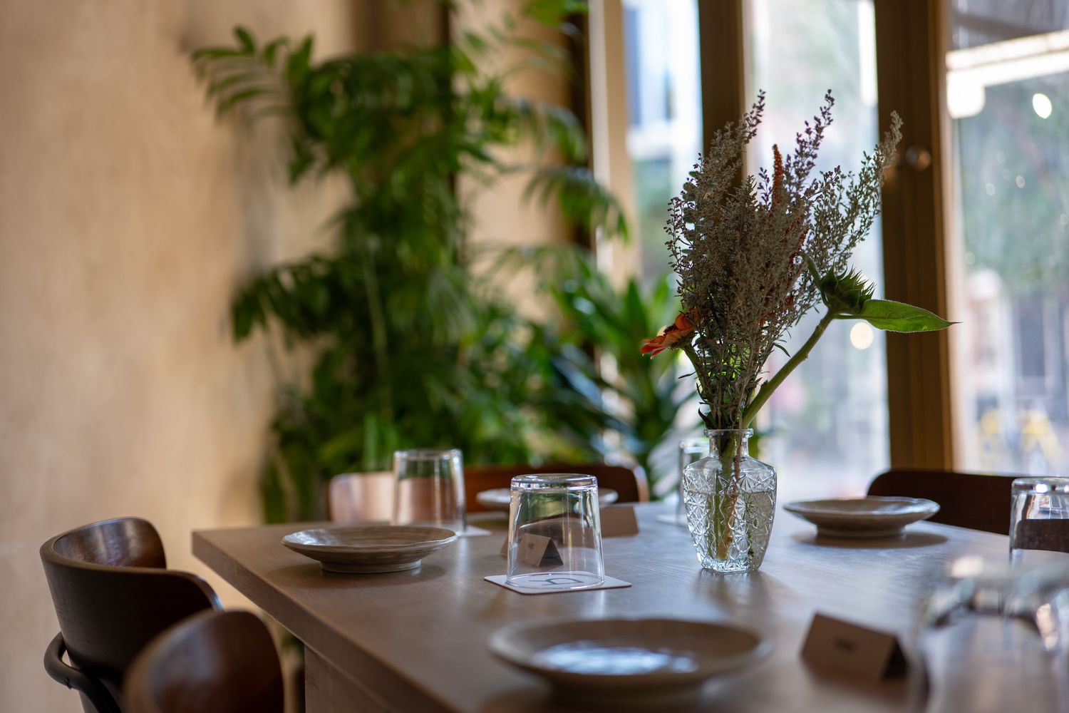 A minimalist restaurant table setting with lavender flowers in a vase, glassware, and natural light from windows.