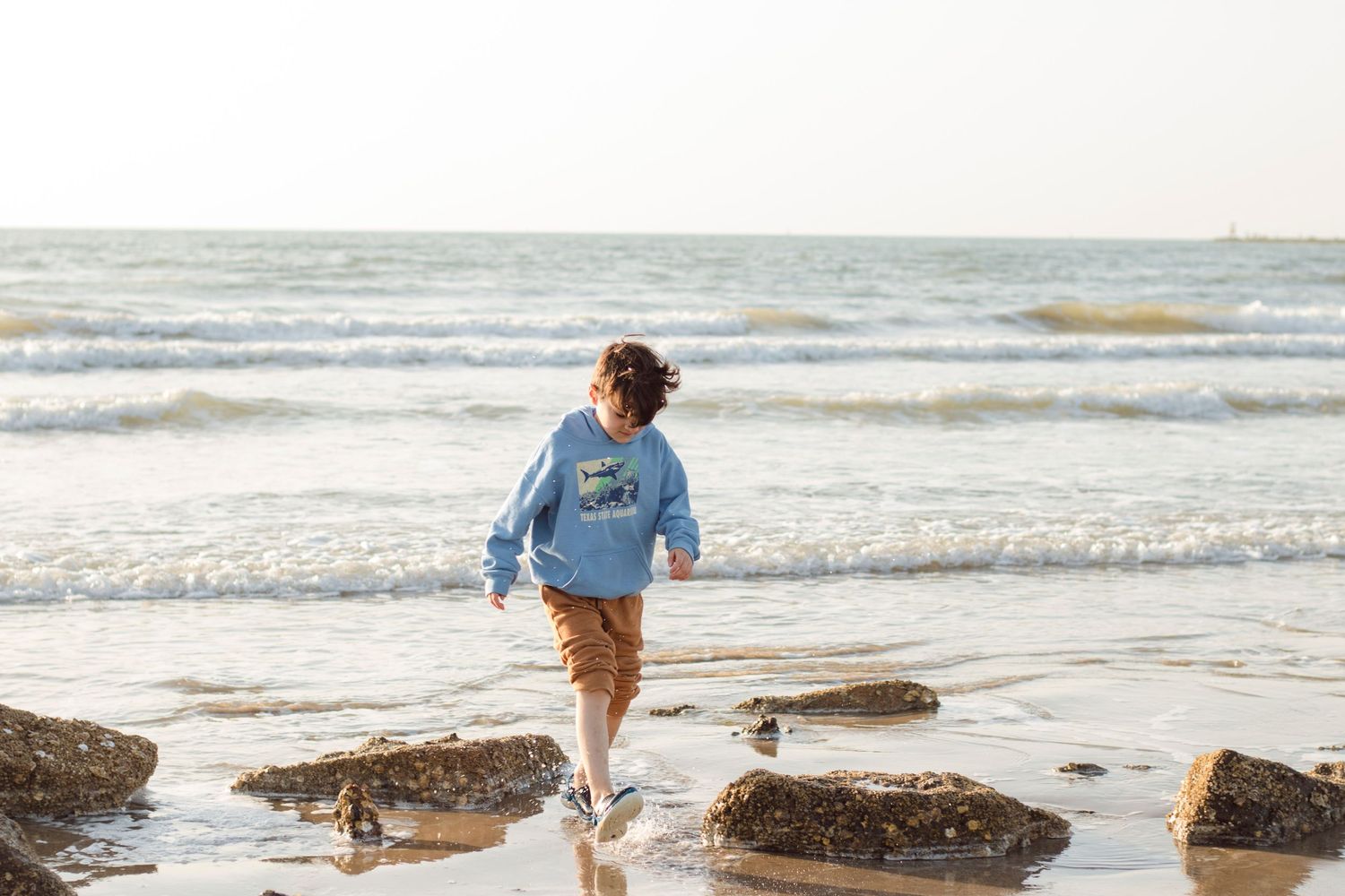 Boy in a blue sweatshirt walks along a rocky beach with crashing waves in the background at sunrise in Surfside Texas