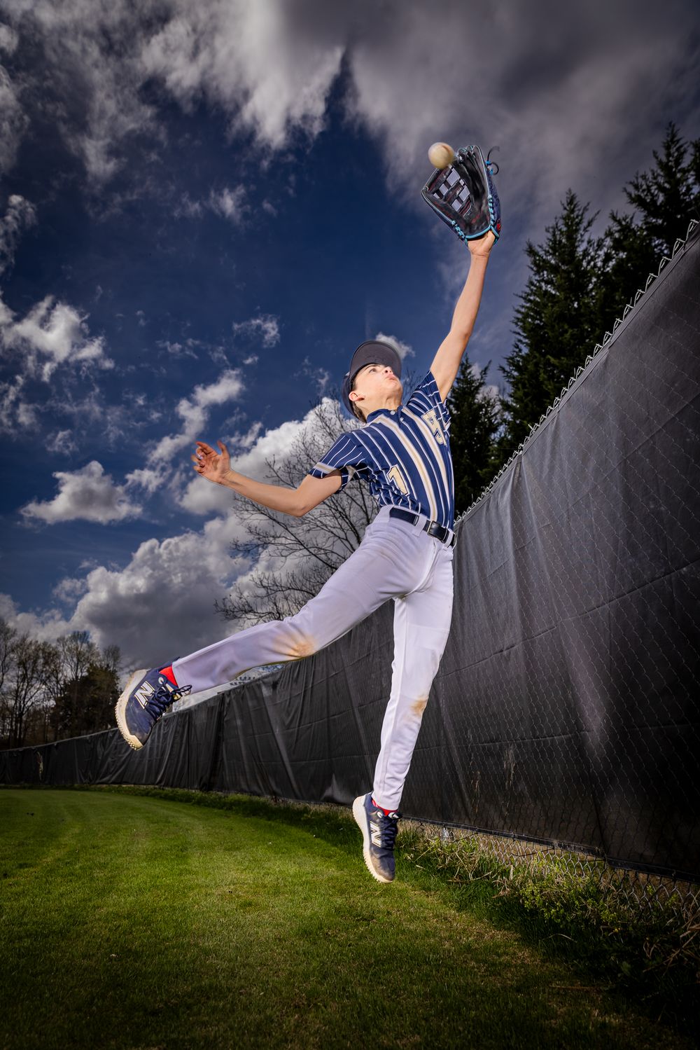Young baseball player leaps near outfield wall, reaching up with glove to catch ball under dramatic cloudy sky.