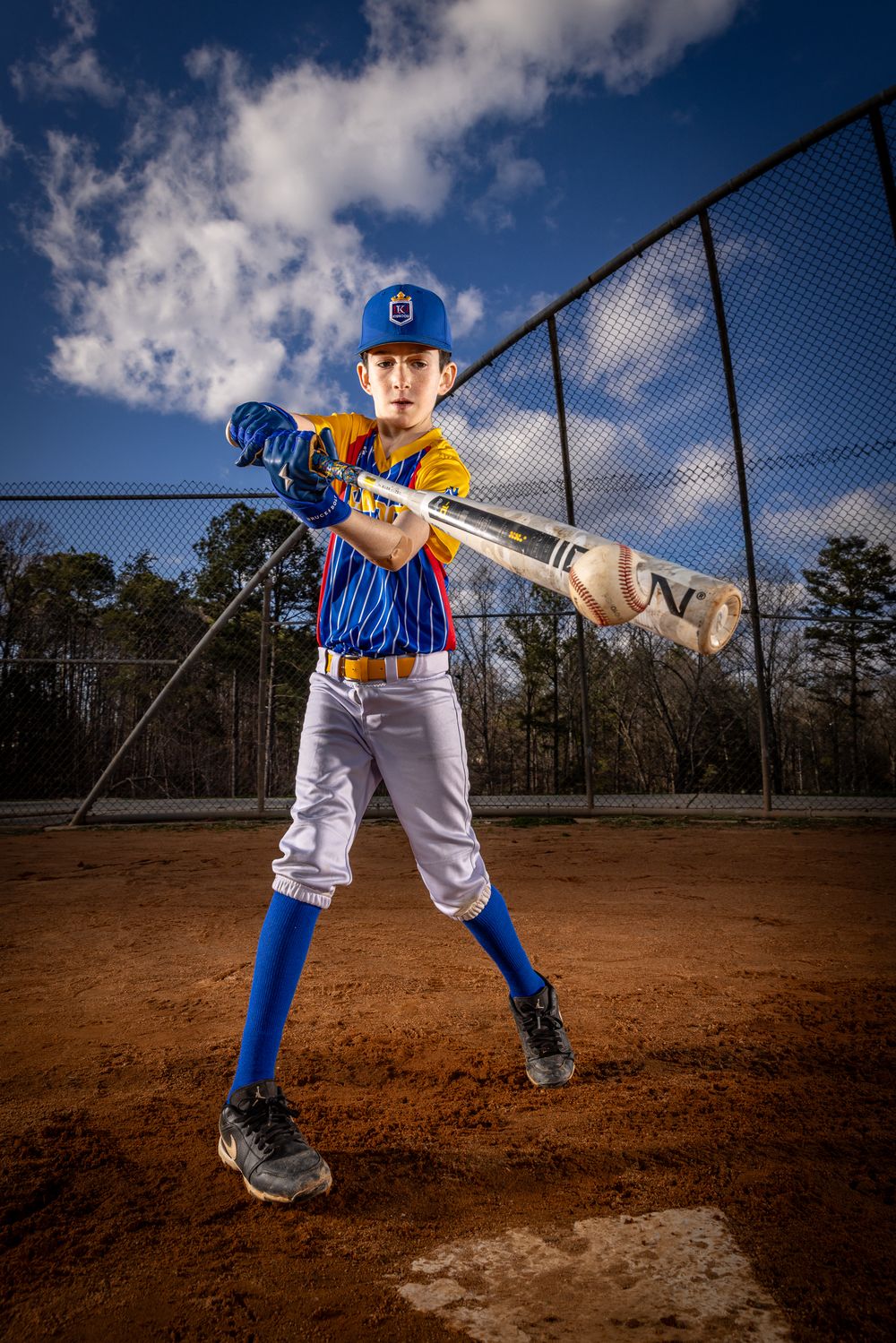Young baseball player in blue uniform holds bat at home plate under dramatic cloudy sky on dirt field.