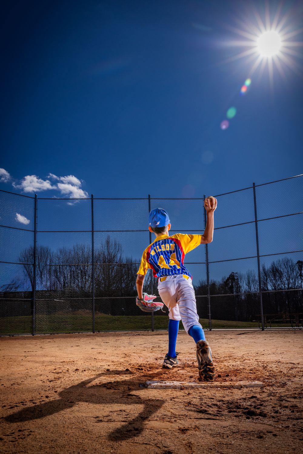 Youth baseball pitcher in yellow and blue uniform winds up to throw on a sunny day at a dirt mound.