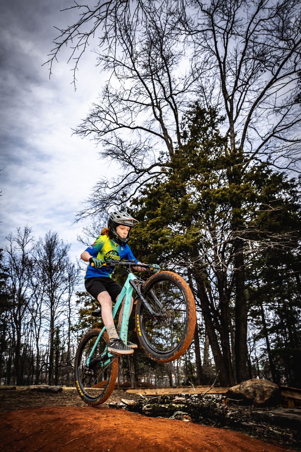 Female mountain biker performing a wheelie on red dirt trail surrounded by bare trees under dramatic cloudy sky.