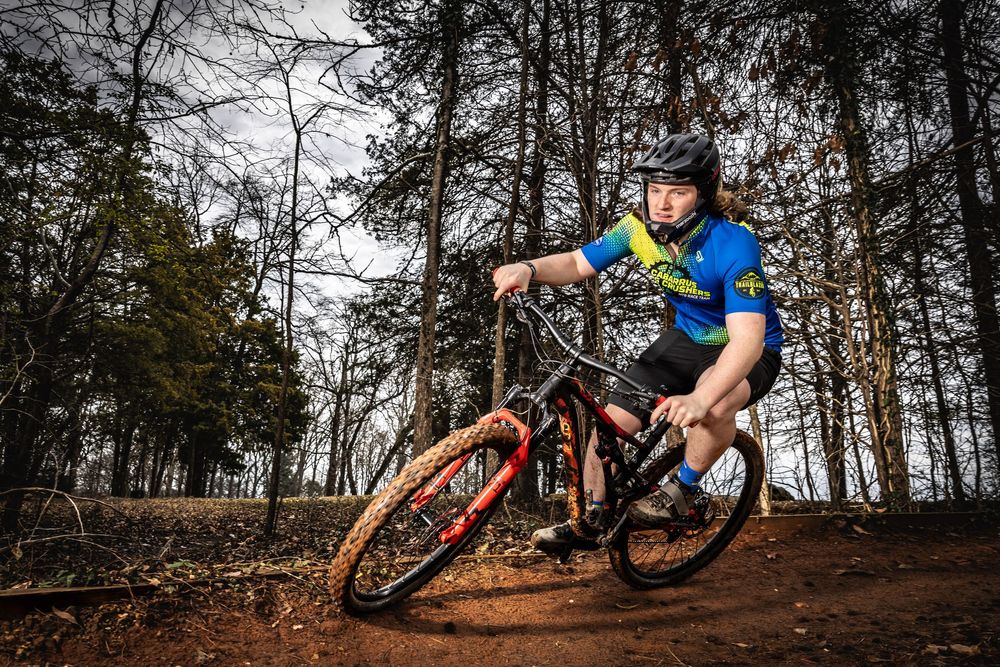 Mountain biker riding red bike on forest trail, wearing blue jersey and helmet, performing sharp turn on dirt path.