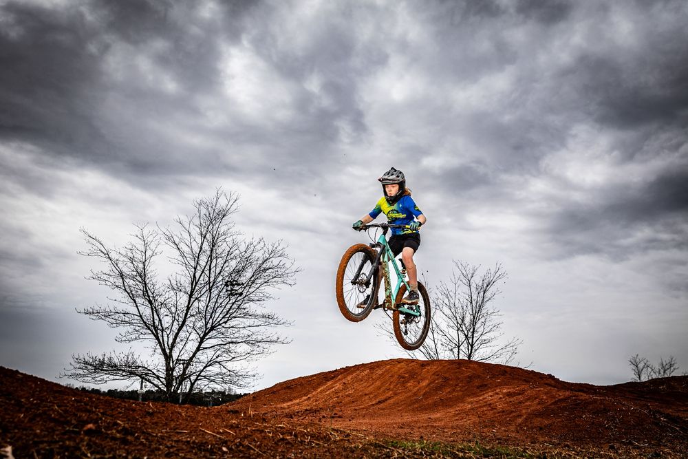 A young mountain biker catches air off a red dirt jump under dramatic cloudy skies with bare trees in the background.