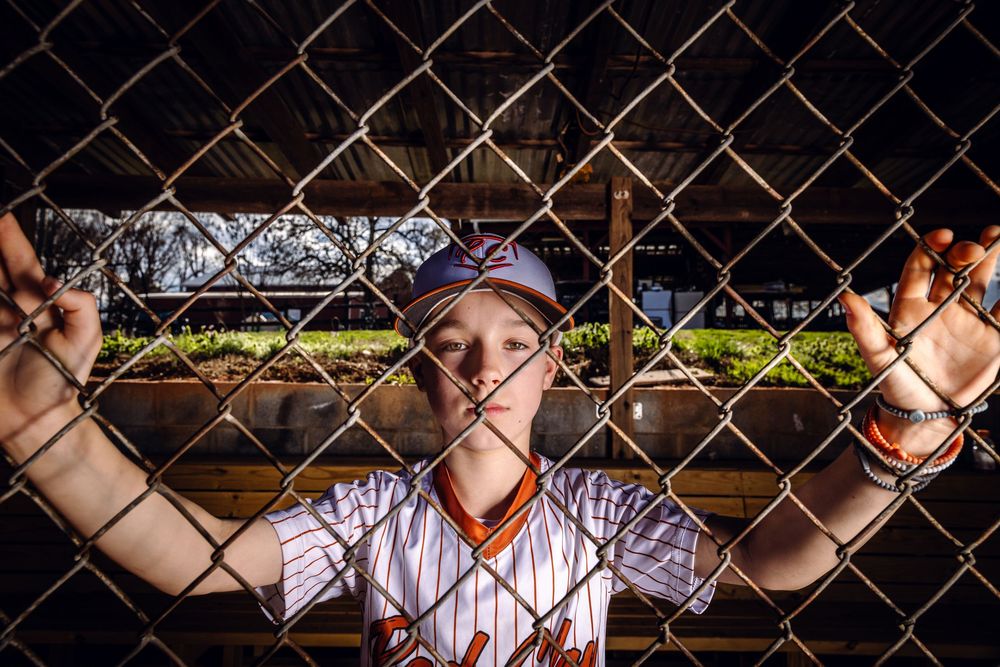 Young baseball player in uniform gripping chain-link fence dugout, dramatic dark lighting on sports field.