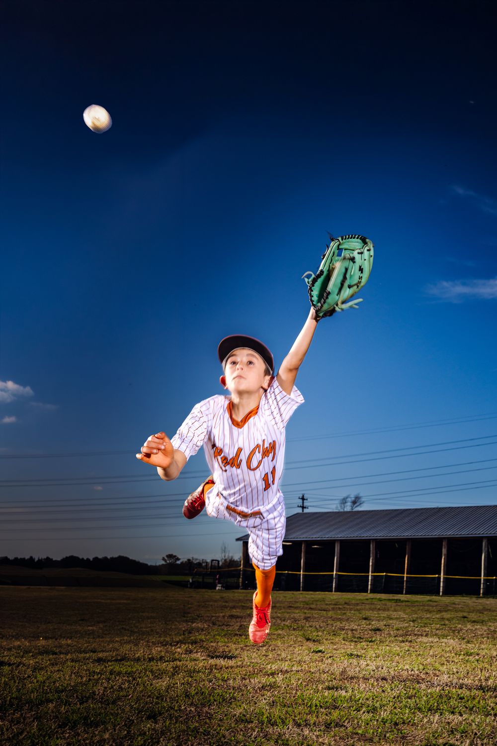 Young baseball player in white pinstripe uniform leaps to catch a ball with green glove under blue sky.
