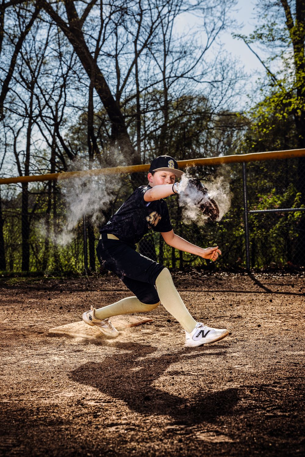 Young baseball player in black uniform slides into base in a cloud of dust at an outdoor dirt field.