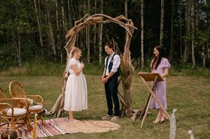 Rustic outdoor wedding ceremony setup with wooden arch, woven baskets and bohemian rugs in a forest clearing.