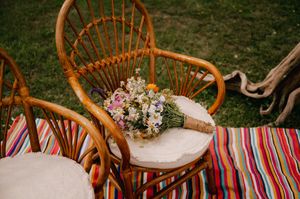 Rustic rattan chair with floral bouquet on striped picnic blanket in outdoor garden setting.
