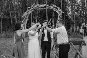 A candid moment during a rustic outdoor wedding celebration beneath a wooden arch in a wooded setting.