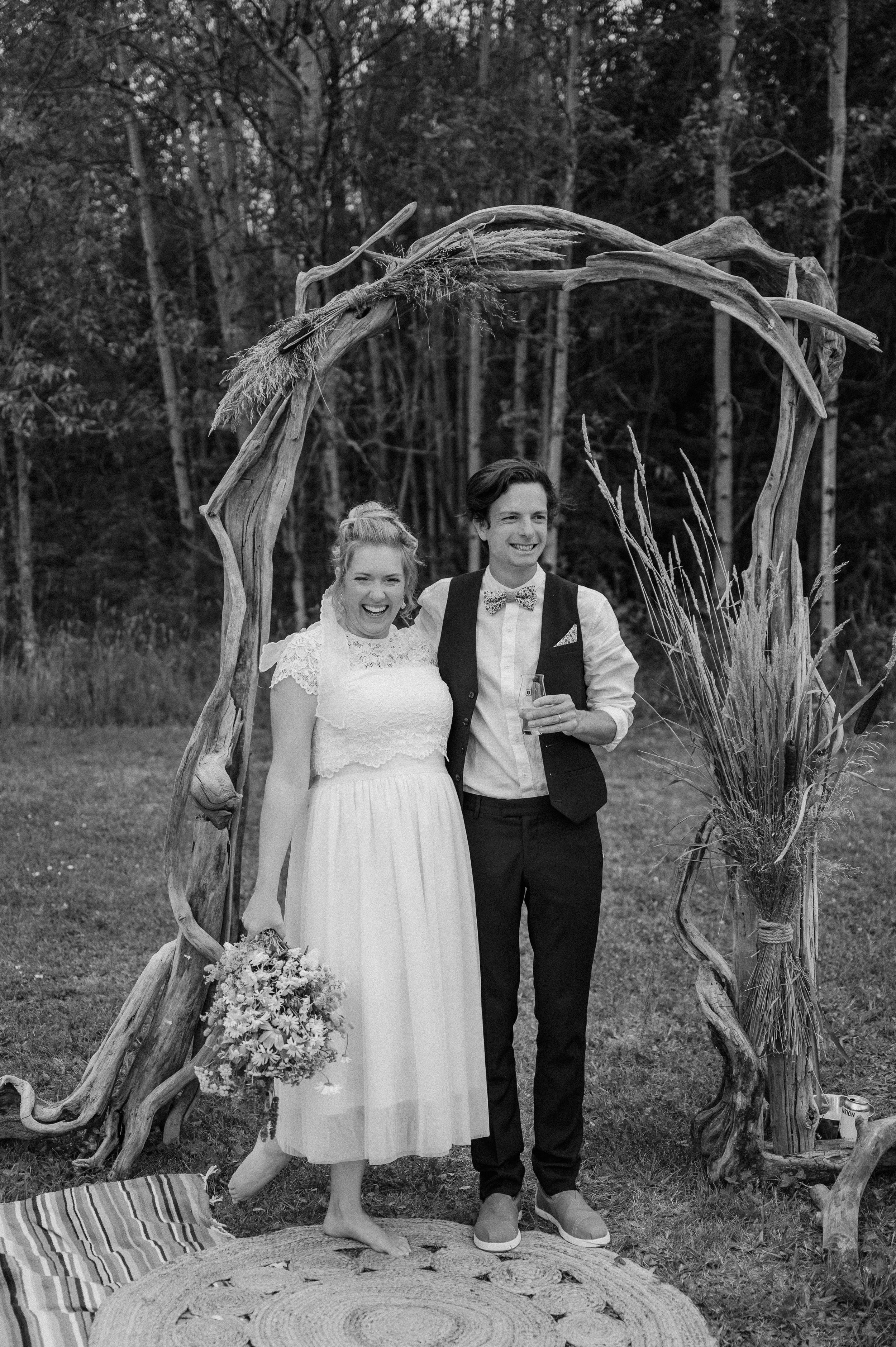 A couple stands beneath a rustic driftwood arch at an outdoor wedding ceremony in black and white.