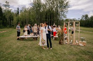 A rustic outdoor wedding ceremony setup in a grassy field surrounded by trees with wooden decor and guests gathered.