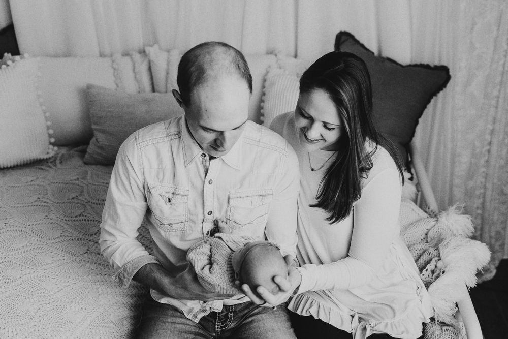 Parents in coordinating white outfits share tender moments with their newborn on a light couch.