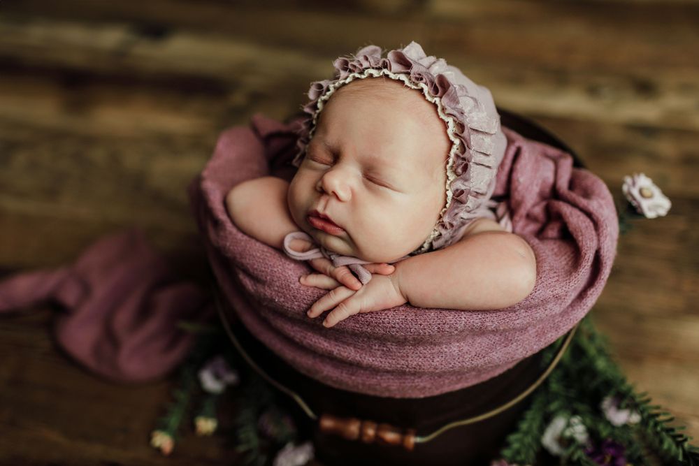 Newborn baby wrapped in mauve fabric wearing a delicate lace bonnet sleeping in wooden basket.