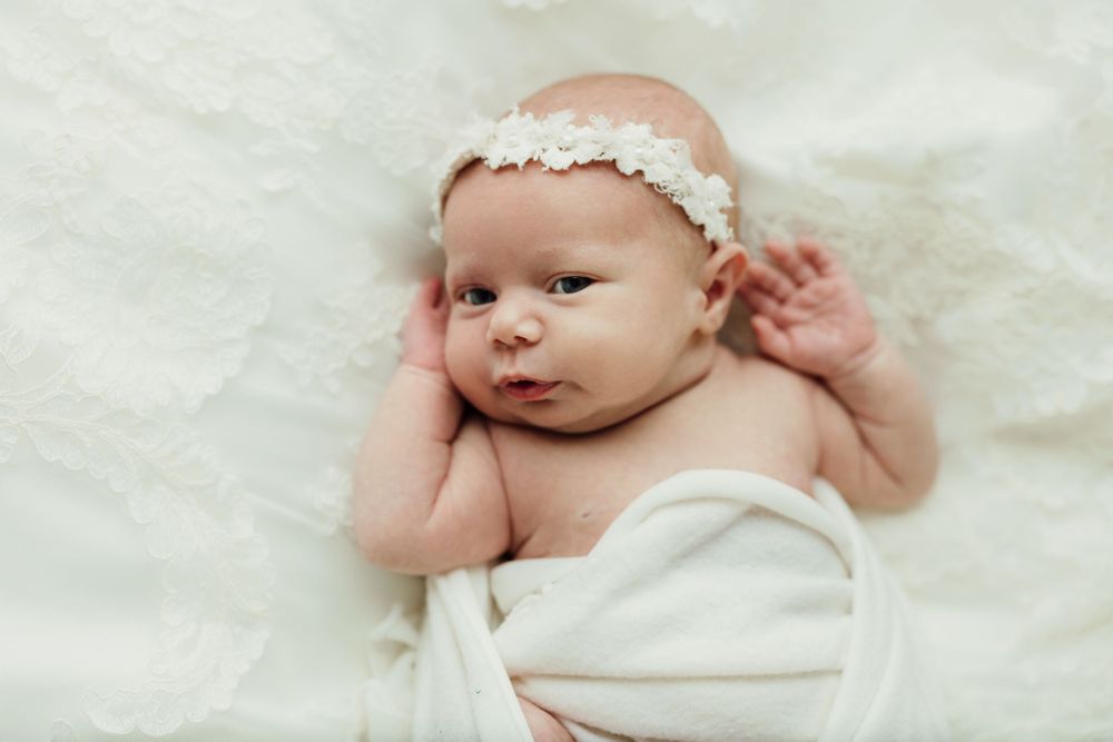 Baby wearing white lace headband lying on cream fabric with arms raised.