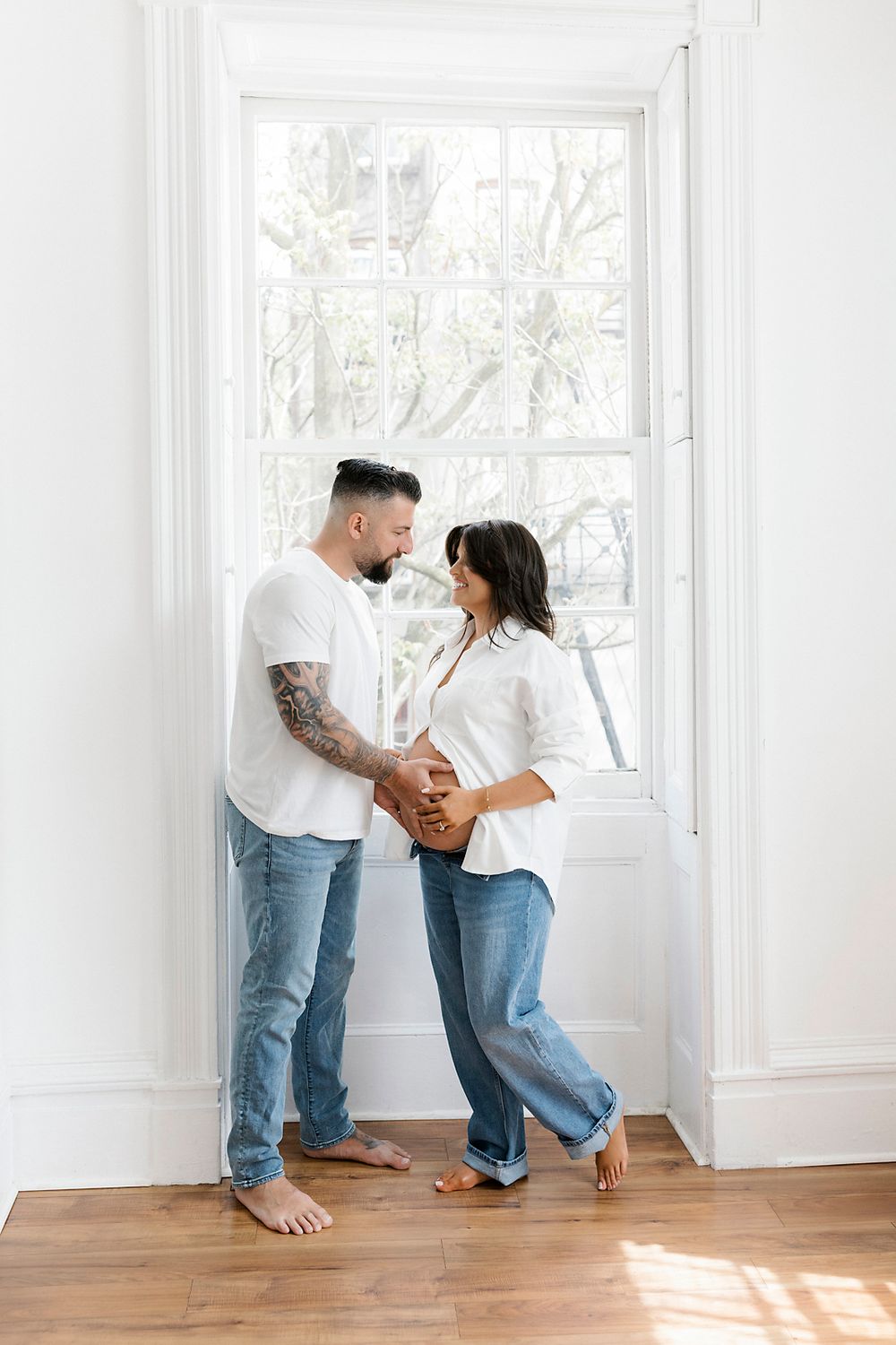 Couple in matching jeans and white shirts share a tender moment by a bright window in their home.