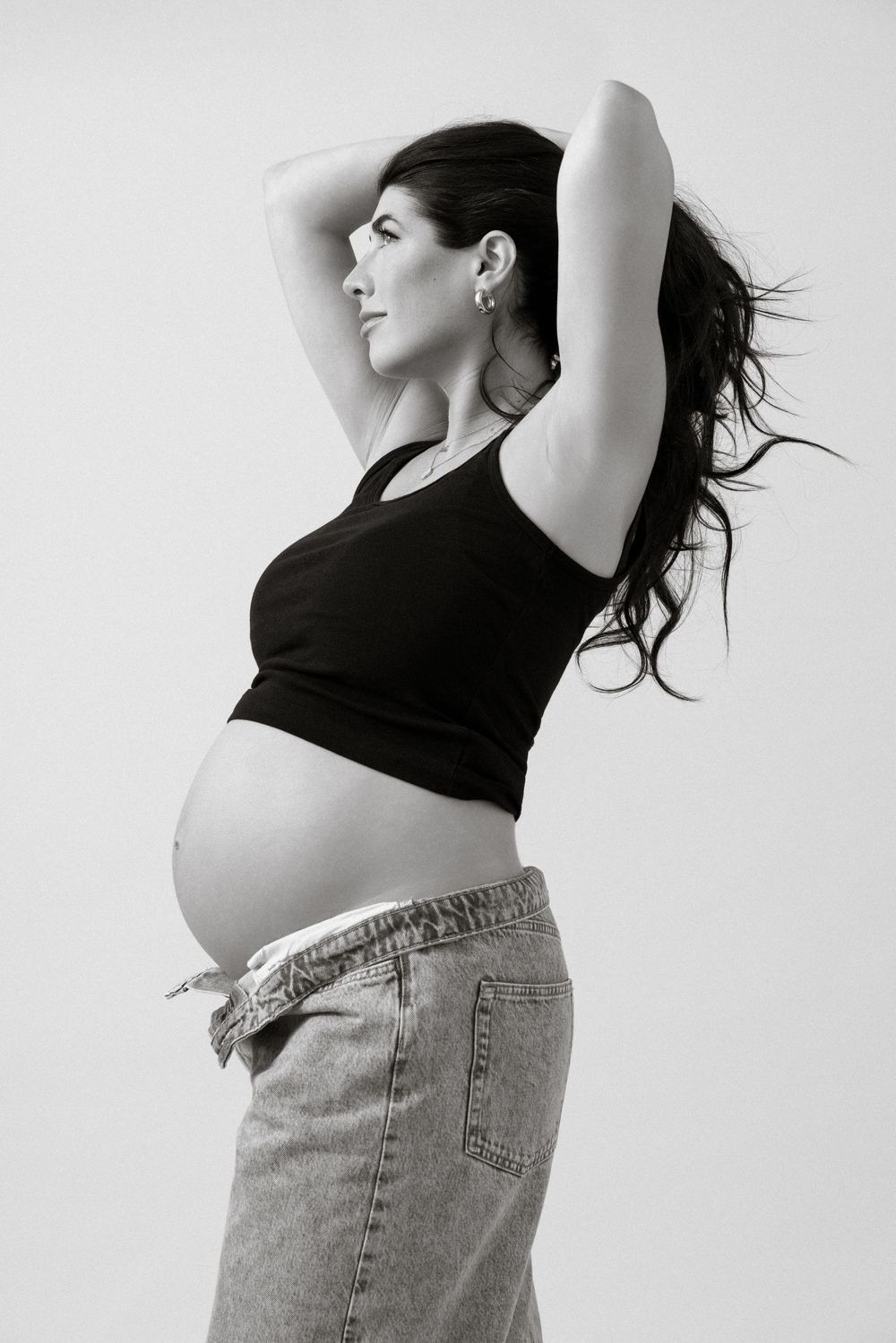 Black and white photo of a pregnant woman in a black tank top and jeans, posing with arms raised.