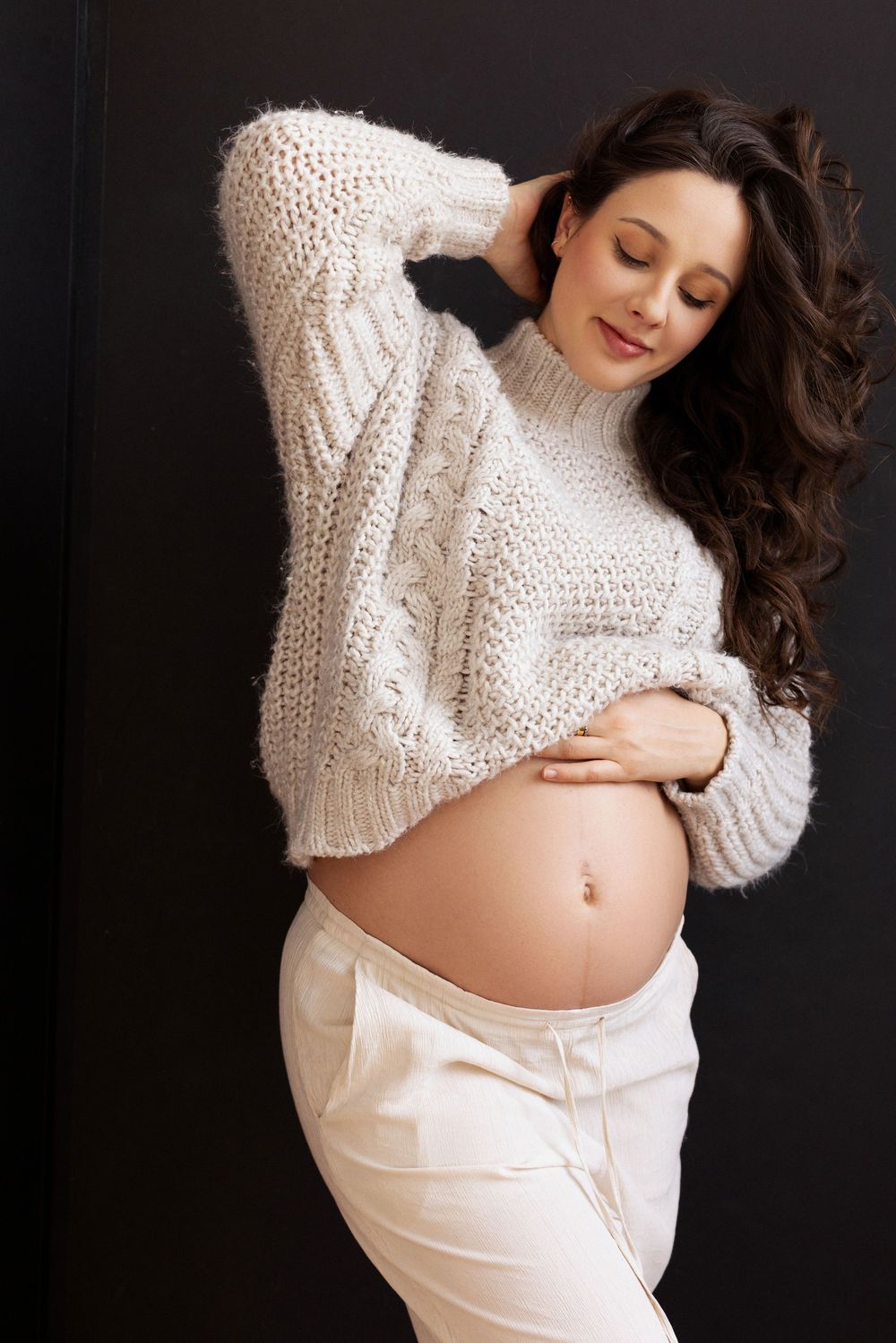 Maternity photo series showing an expectant mother in a white sweater and white fabric against dark background.