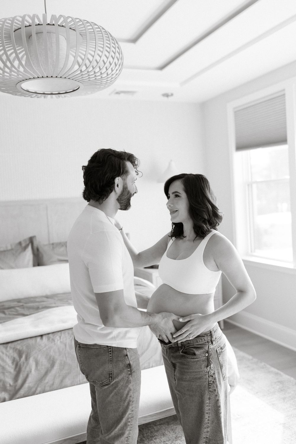 A couple in casual attire dances together in an empty white room with natural lighting streaming through windows.