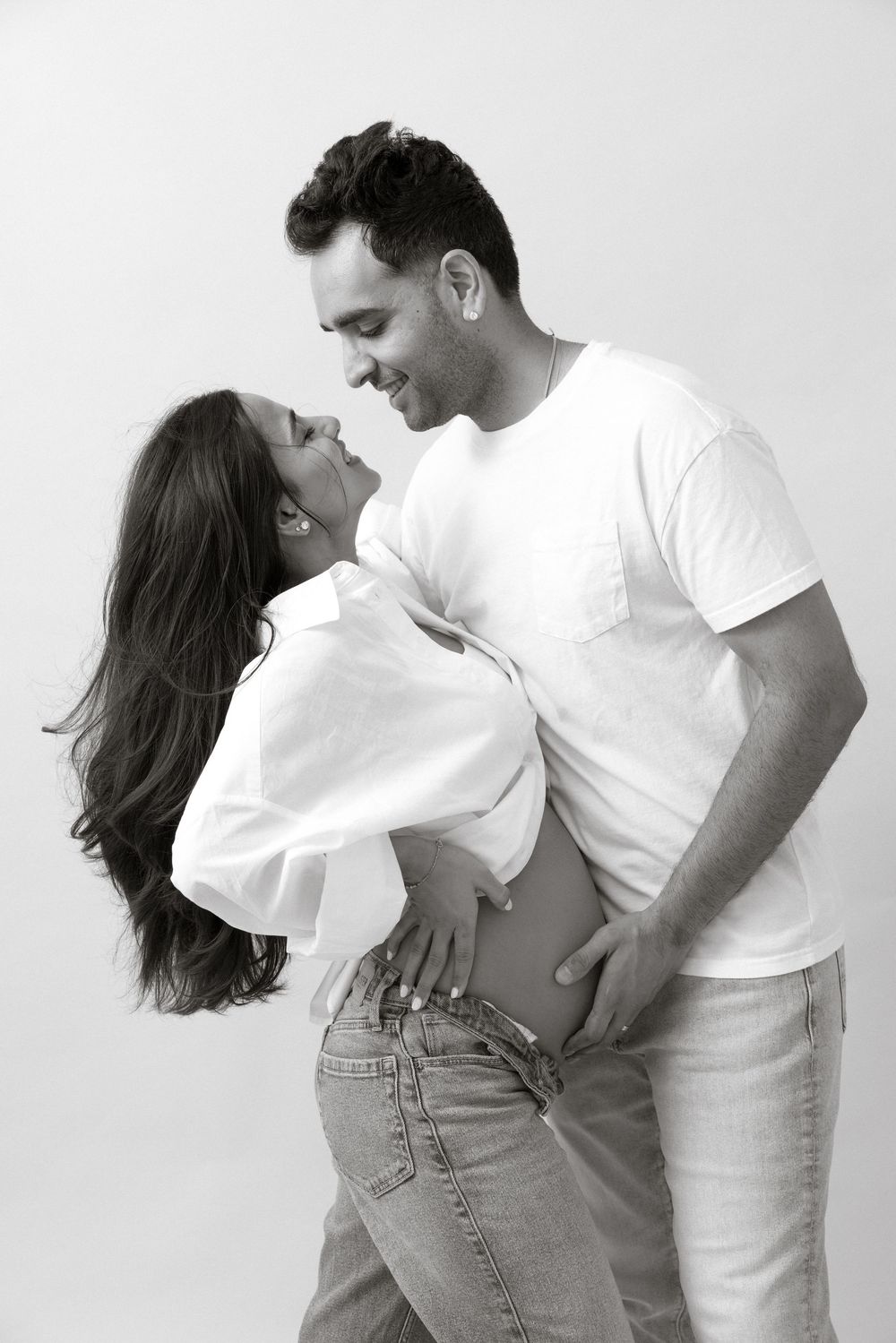 Black and white photo of a smiling pregnant couple embracing, showing baby bump, wearing white shirts and jeans.