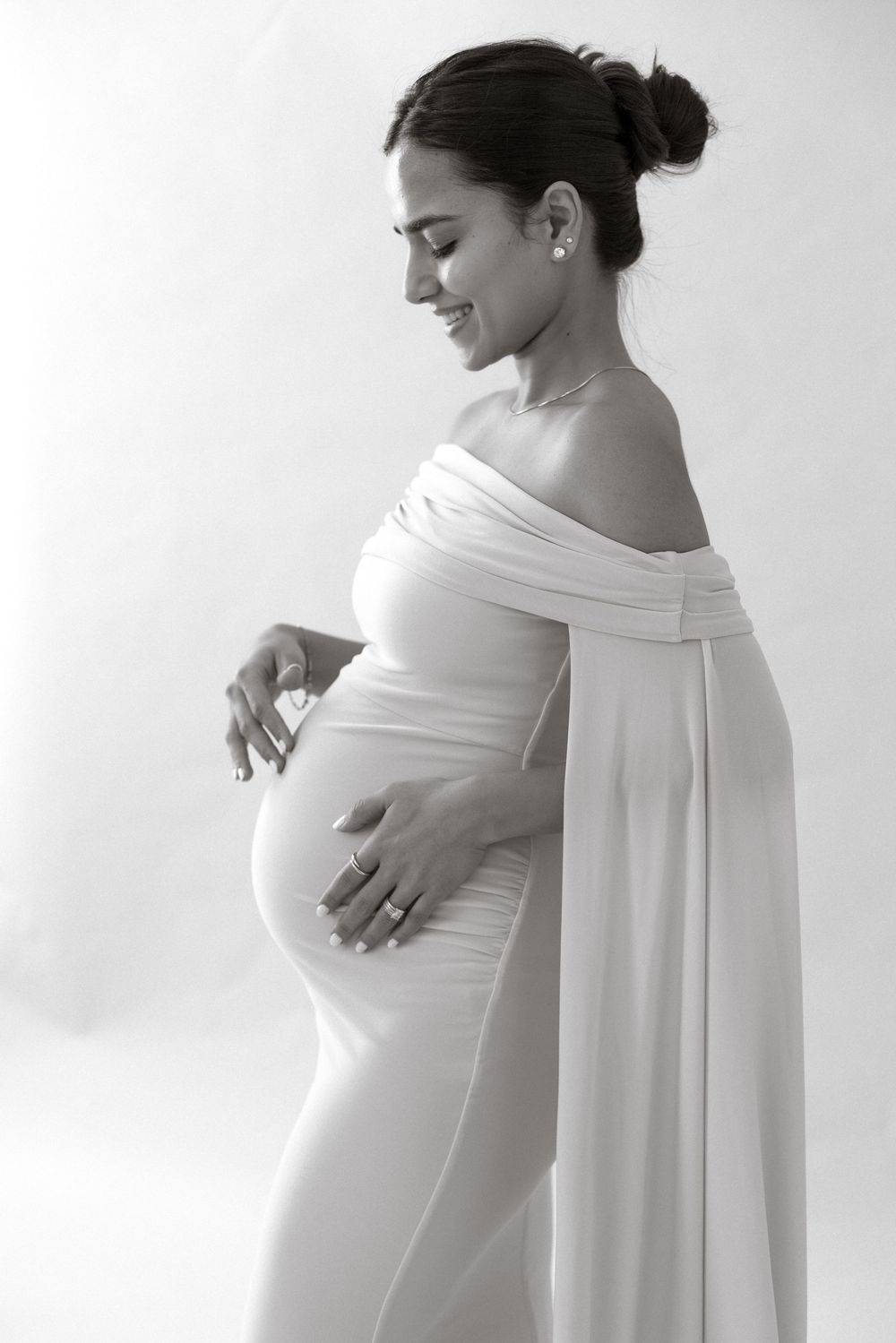 Black and white maternity photo of a smiling pregnant woman in a white off-shoulder gown cradling her baby bump.