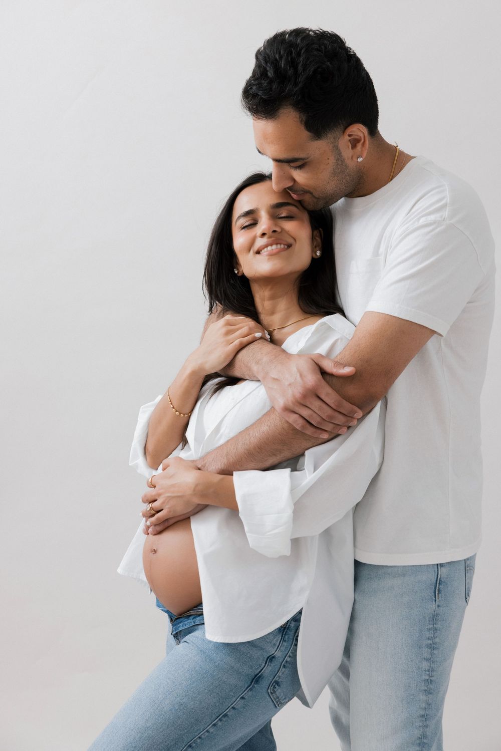 A man tenderly embraces a smiling pregnant woman from behind, both wearing white tops and jeans against a gray background.