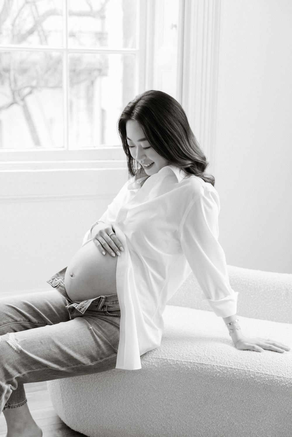 Black and white maternity photo of a pregnant woman in white shirt and jeans, sitting on a sofa near a bright window.