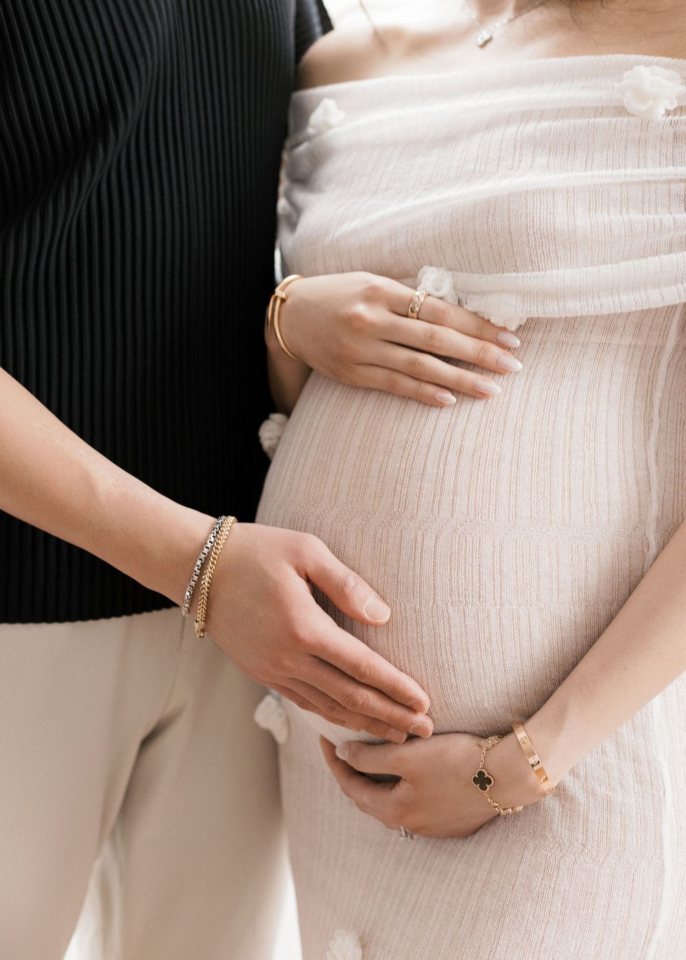 Pregnant woman in white dress with partner's hands cradling baby bump, wearing gold bracelets and rings.