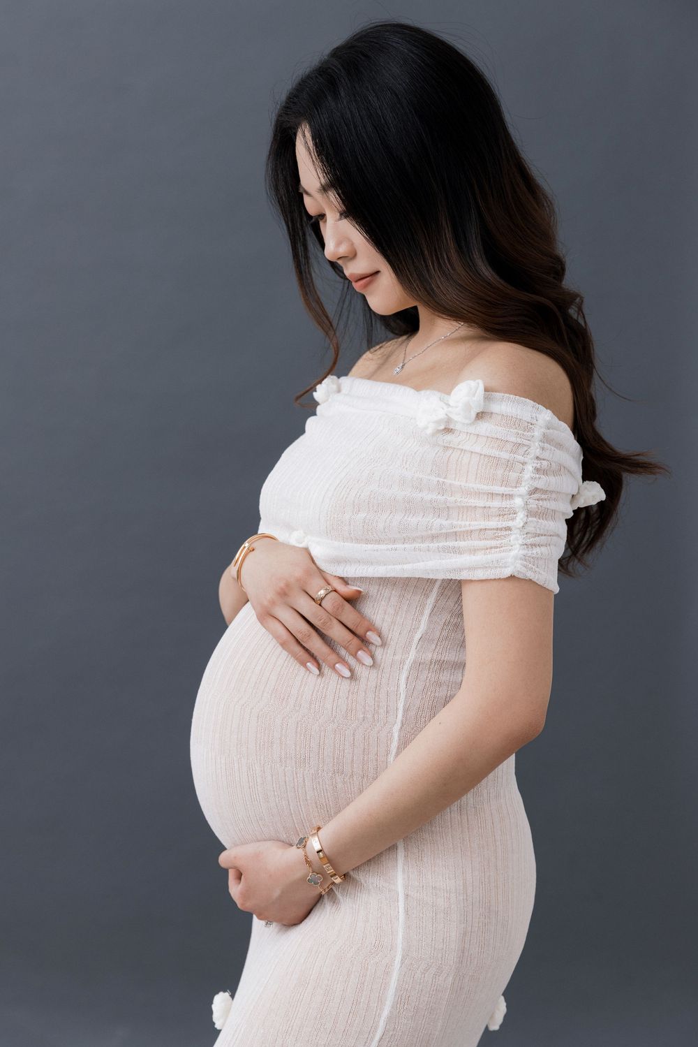 Pregnant woman in white off-shoulder dress cradling baby bump, wearing gold bracelets, against gray background.