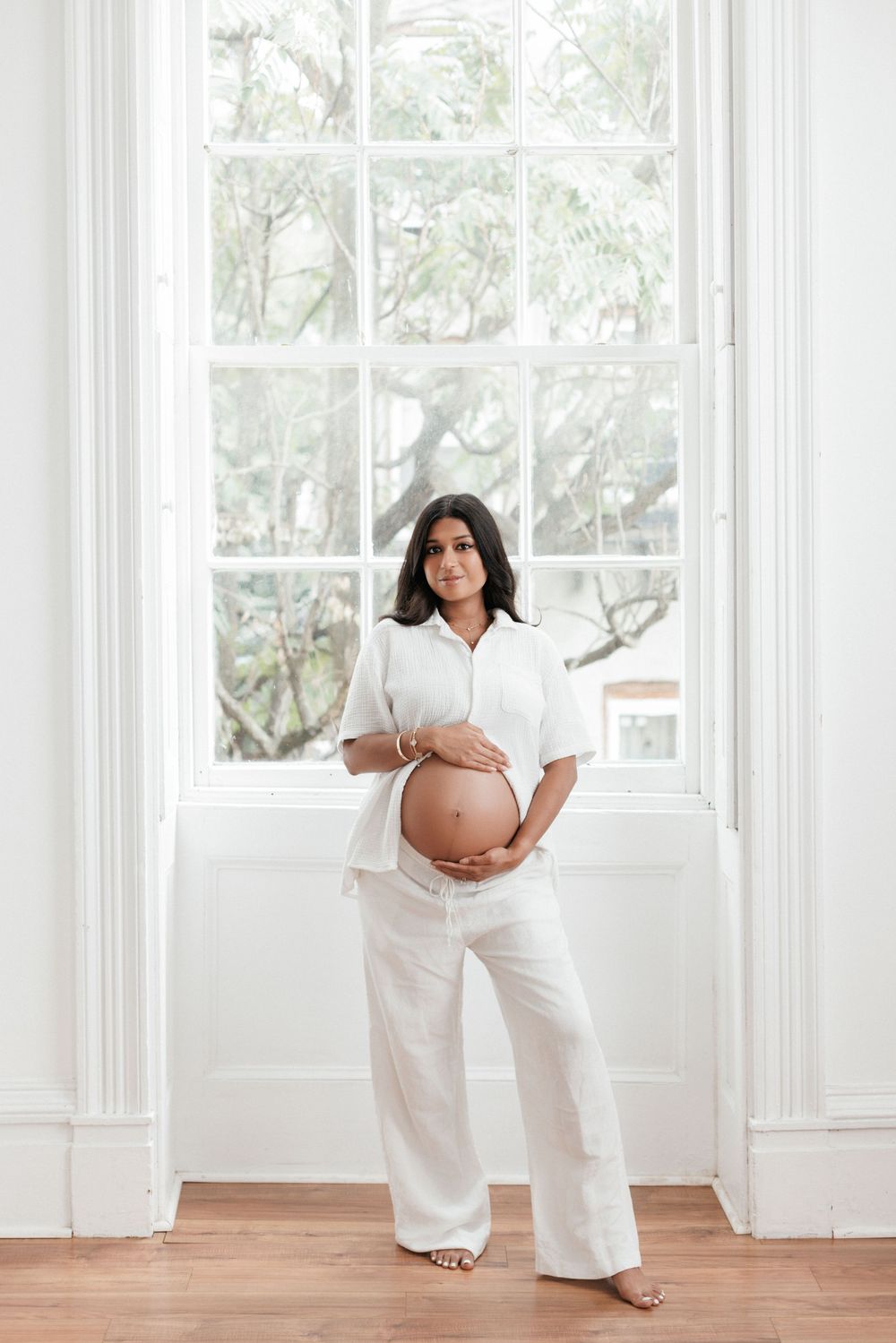Pregnant woman in white outfit cradling baby bump, standing in front of large window with natural light.