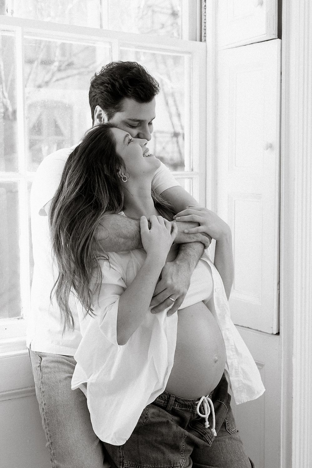 Black and white photo of a romantic couple embracing and kissing passionately by a bright window.