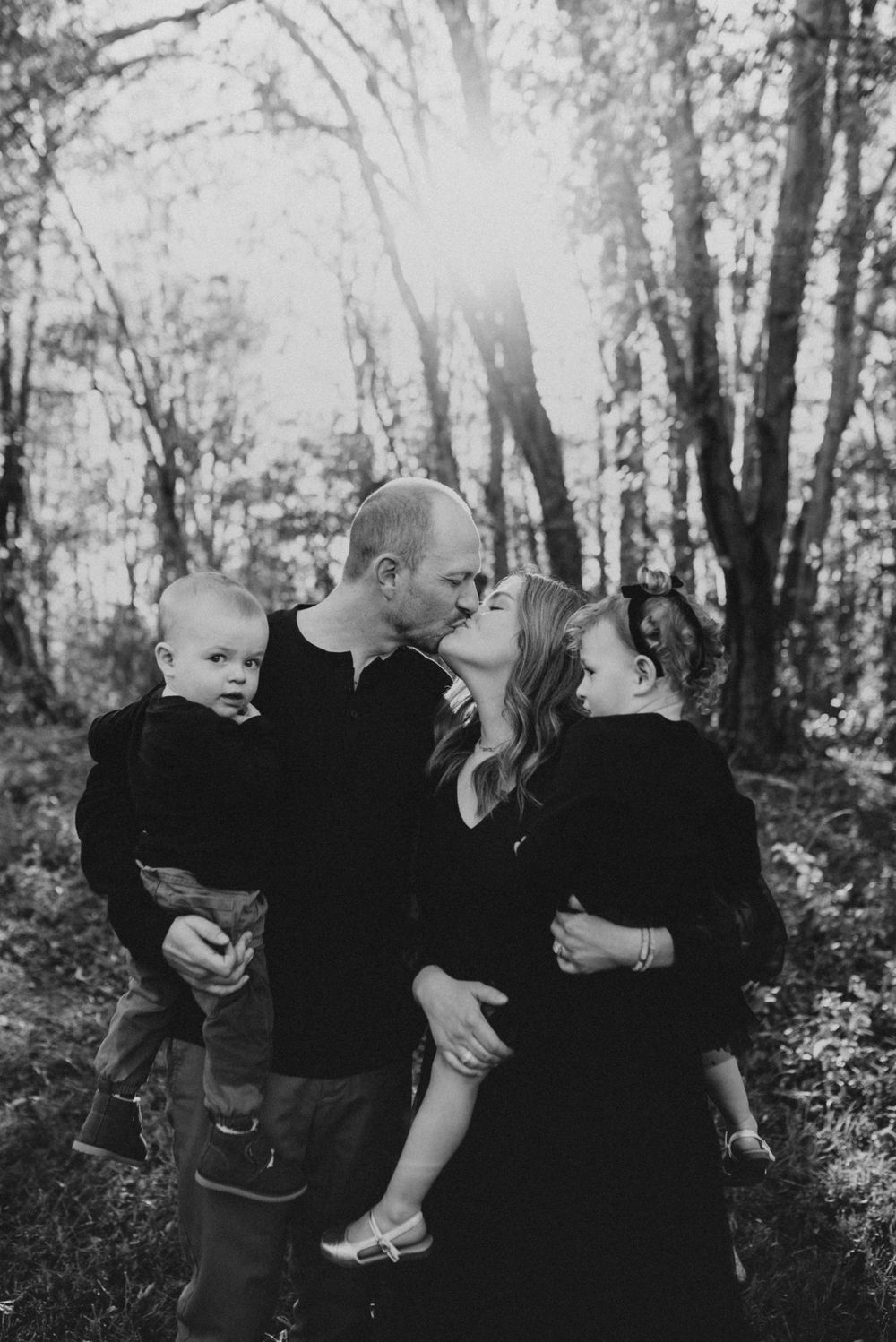 A backlit black and white family portrait in a forest setting with sunlight streaming through trees.