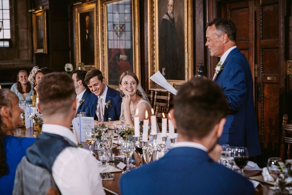 Wedding guests listen attentively to a speech during an elegant reception in a wood-paneled dining hall with ornate portraits Balliol