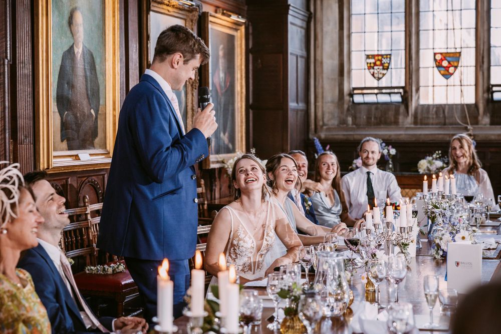 Guests react with laughter during wedding speeches in an elegant hall with historic portraits and stained glass at Balliol College, Oxford