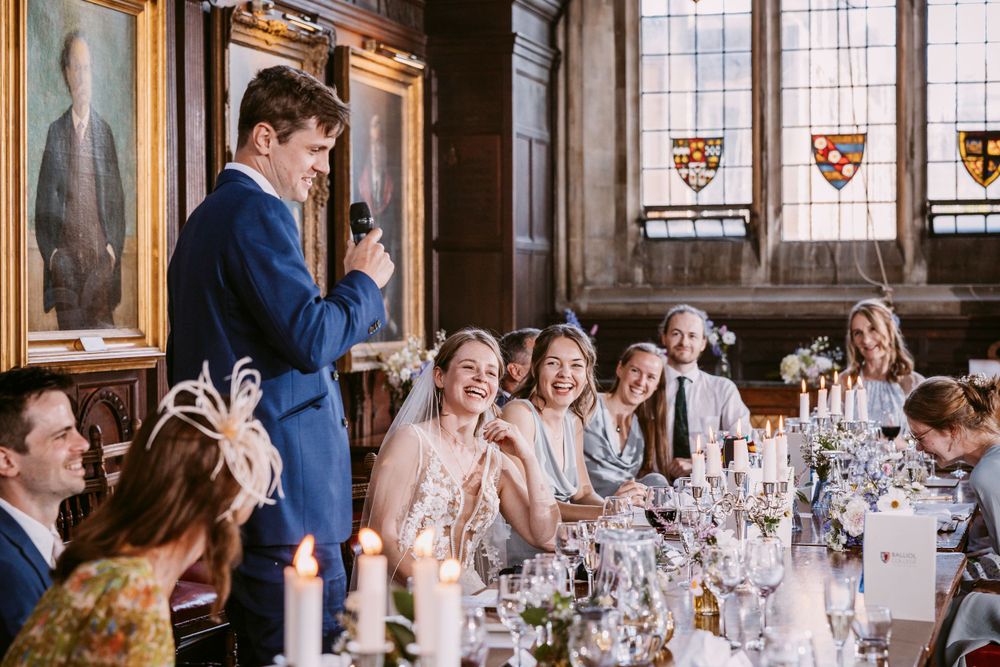Groom delivers a toast during a wedding reception in a grand dining hall with stained glass windows at Balliol College, Oxford