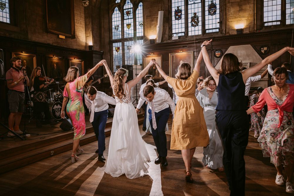Wedding guests dance joyfully in a circle during reception in historic hall with stained glass windows at Balliol College, Oxford