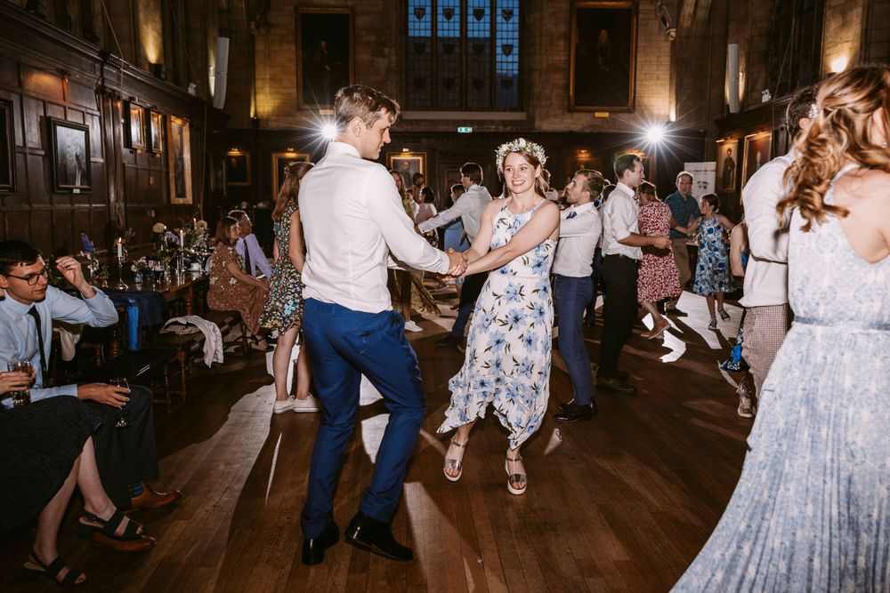 Wedding guests dancing ceilidh energetically in a grand hall with wood paneling and decorative banners at Balliol College, Oxford