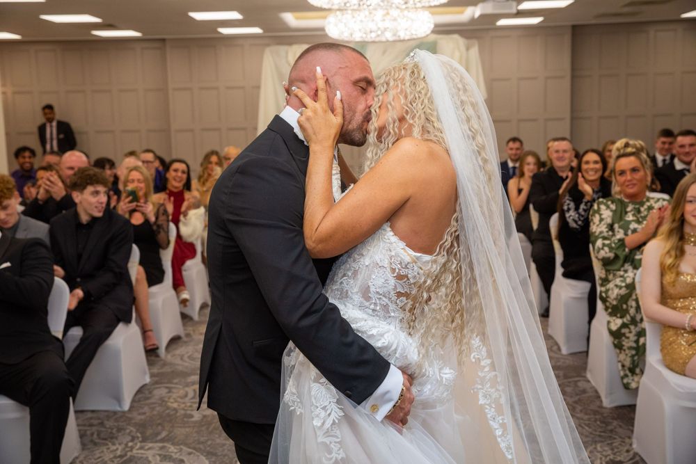 Bride and groom share their first kiss at the wedding altar surrounded by smiling guests in an elegant venue.