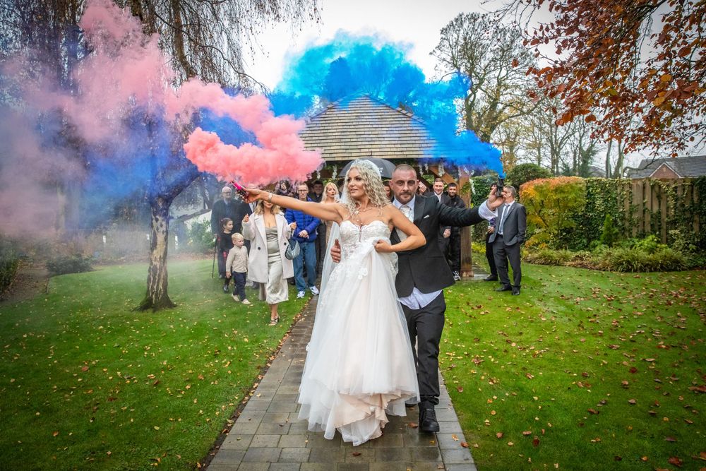 Newlyweds celebrate with colorful pink and blue smoke bombs during outdoor wedding exit in autumn garden.