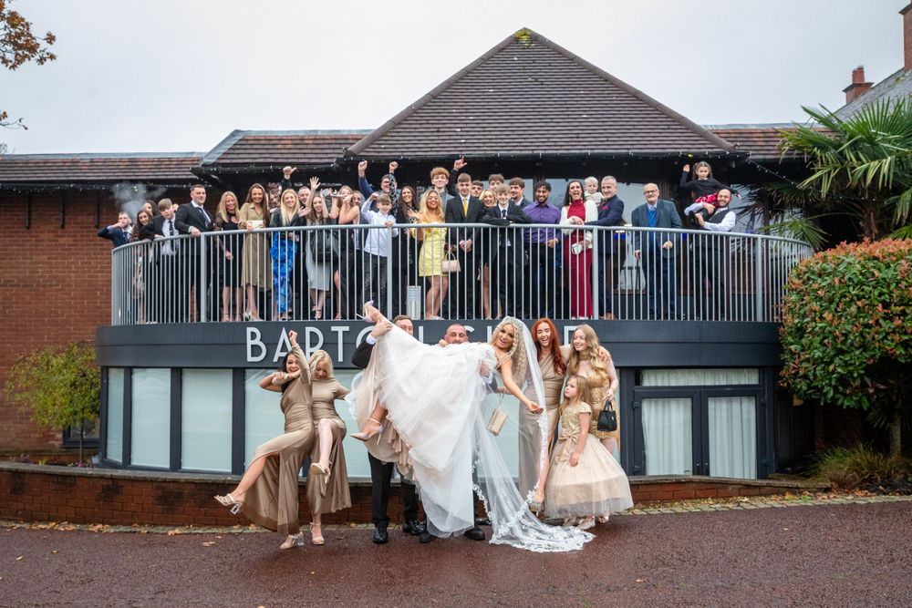 Wedding party poses playfully outside venue as guests cheer from balcony above on a cloudy day.