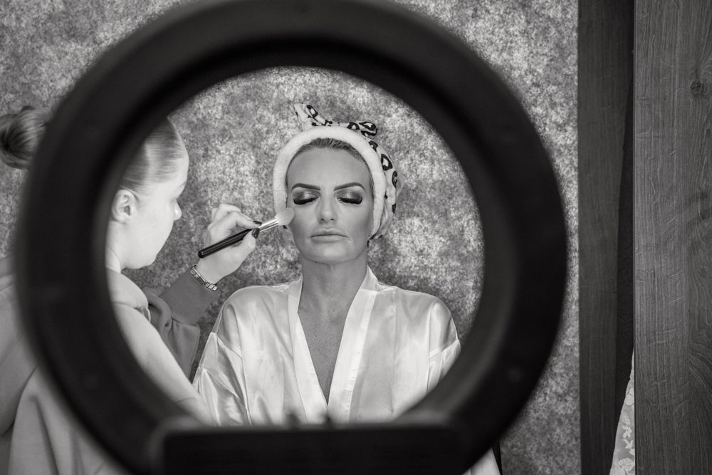 Black and white photo of bride having makeup applied reflected in a ring light mirror.