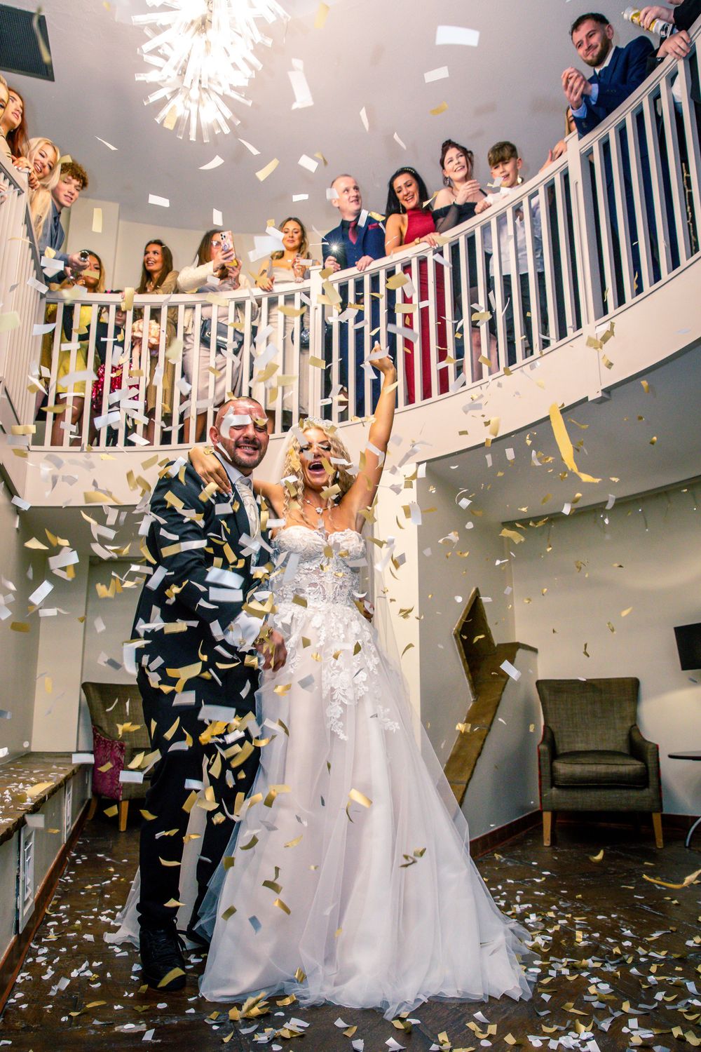 Bride and groom celebrate with golden confetti shower indoors as wedding guests watch from spiral staircase.