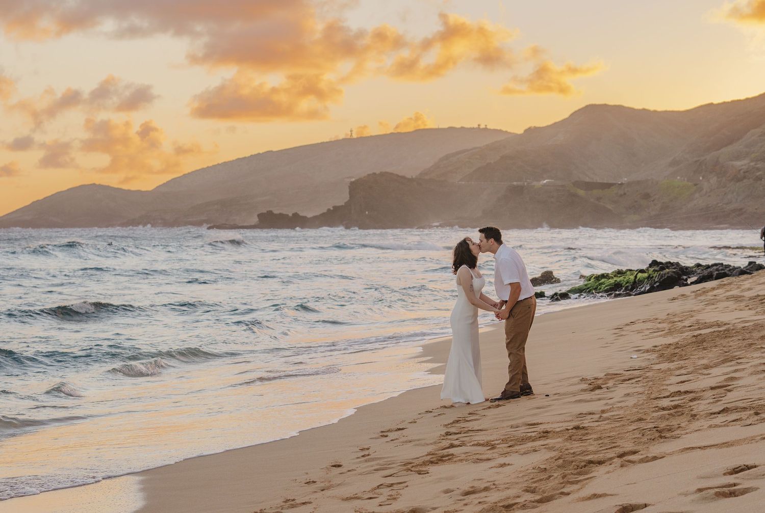 Two people share a romantic sunset moment on a beach with mountains and crashing waves in Hawaii.