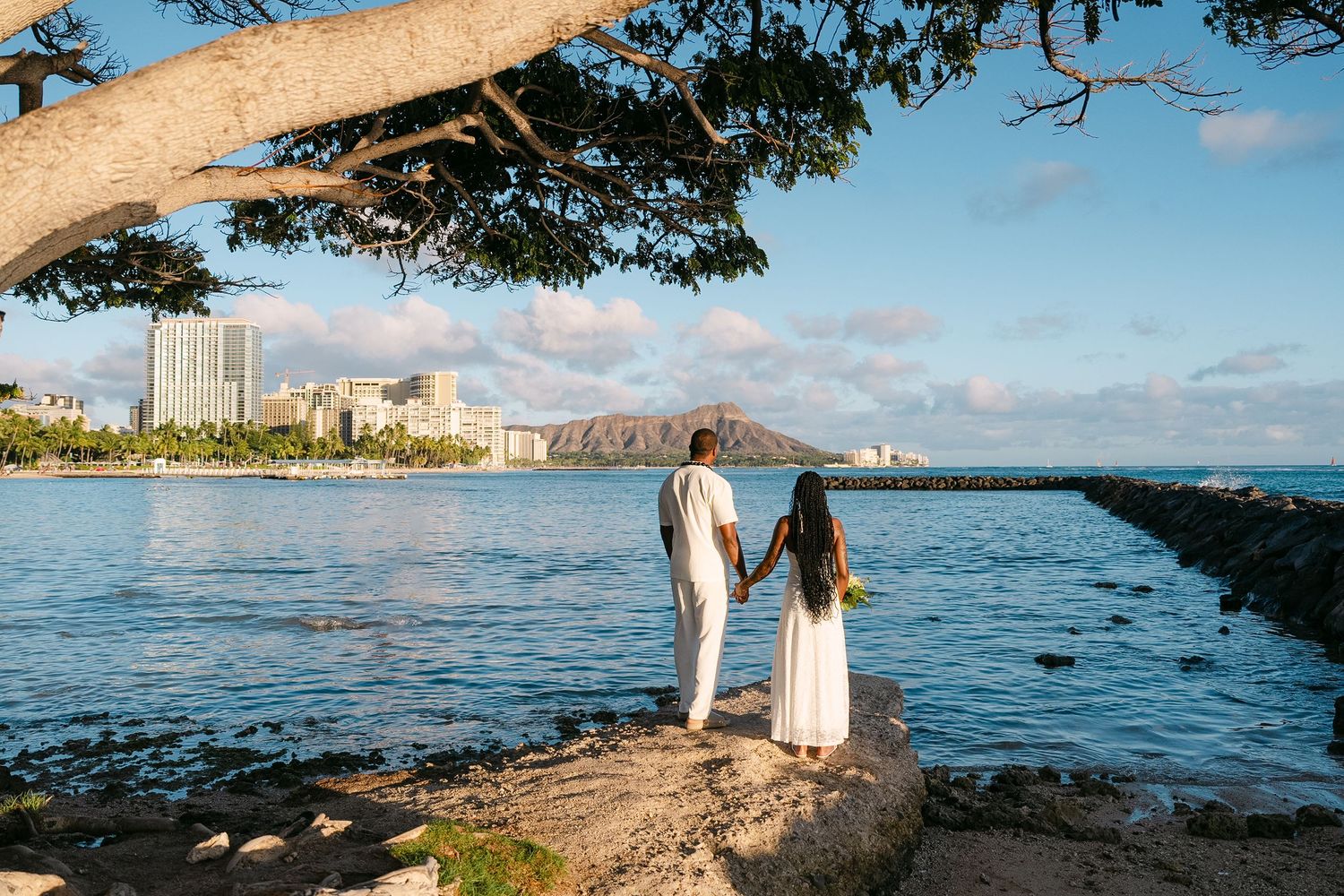 A couple stands on a rocky shoreline overlooking Waikiki Beach and Diamond Head in Honolulu, Hawaii at sunset.