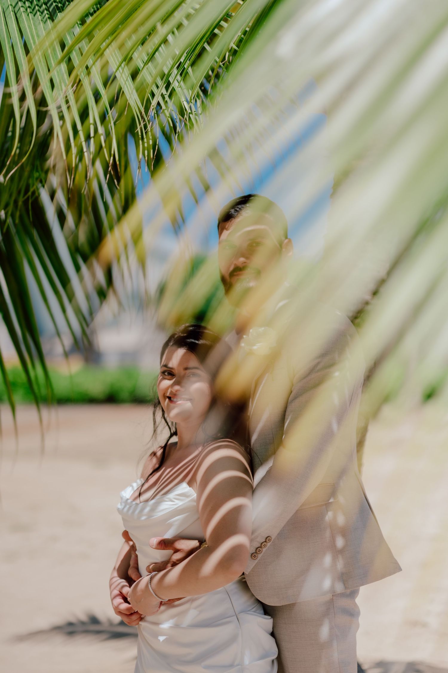 Waialae beach Couple photoshoot A woman in a wedding dress swaying palm fronds casting dappled shadows on a sunny beach.