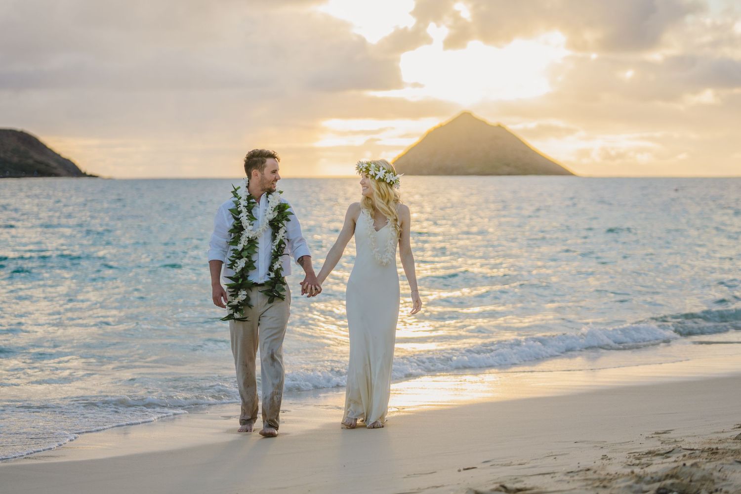 A couple in wedding attire walks along a scenic beach at sunset with mountains and ocean in the background.