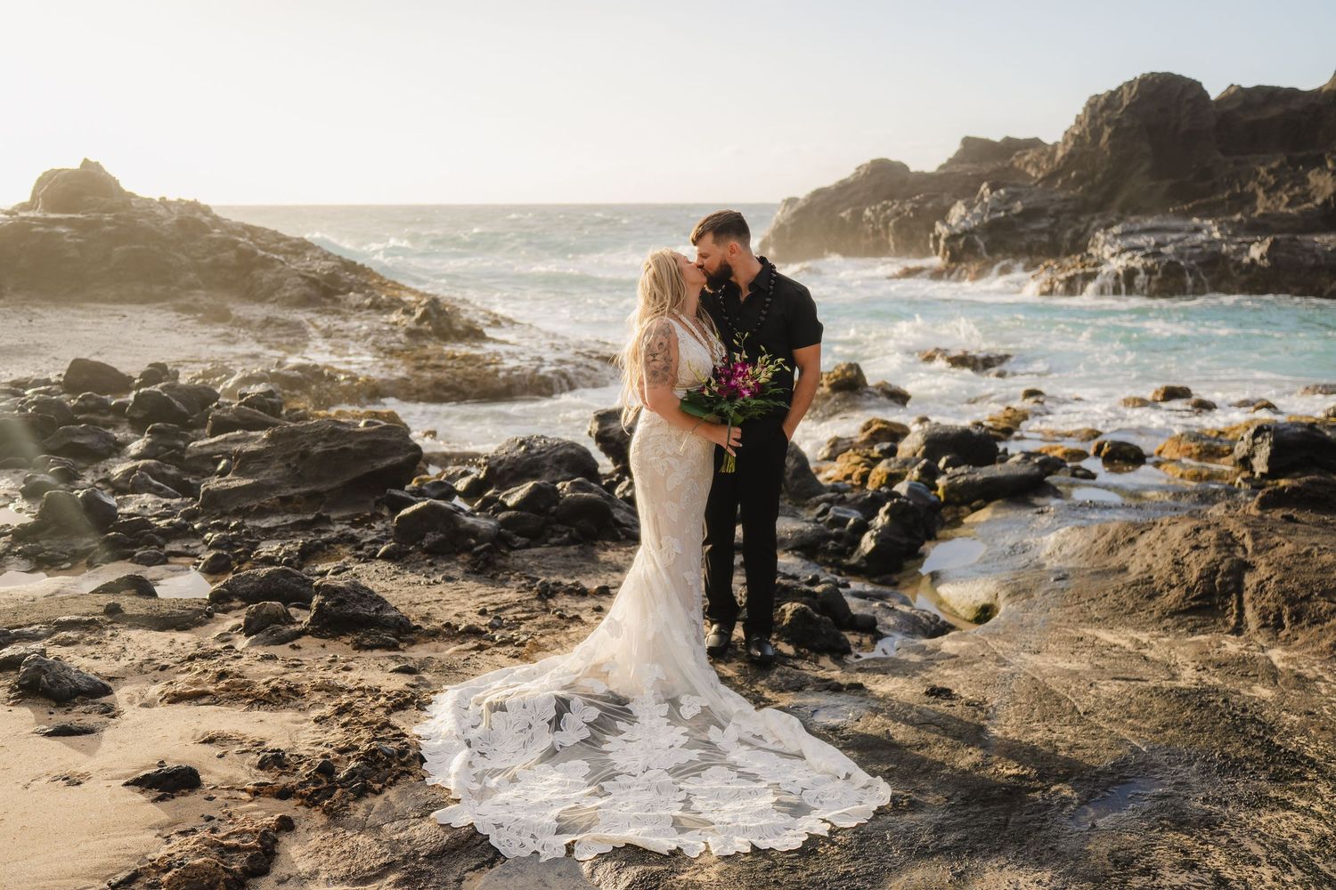 Couple shares a romantic kiss in wedding attire on rocky oceanside beach with crashing waves at sunset.