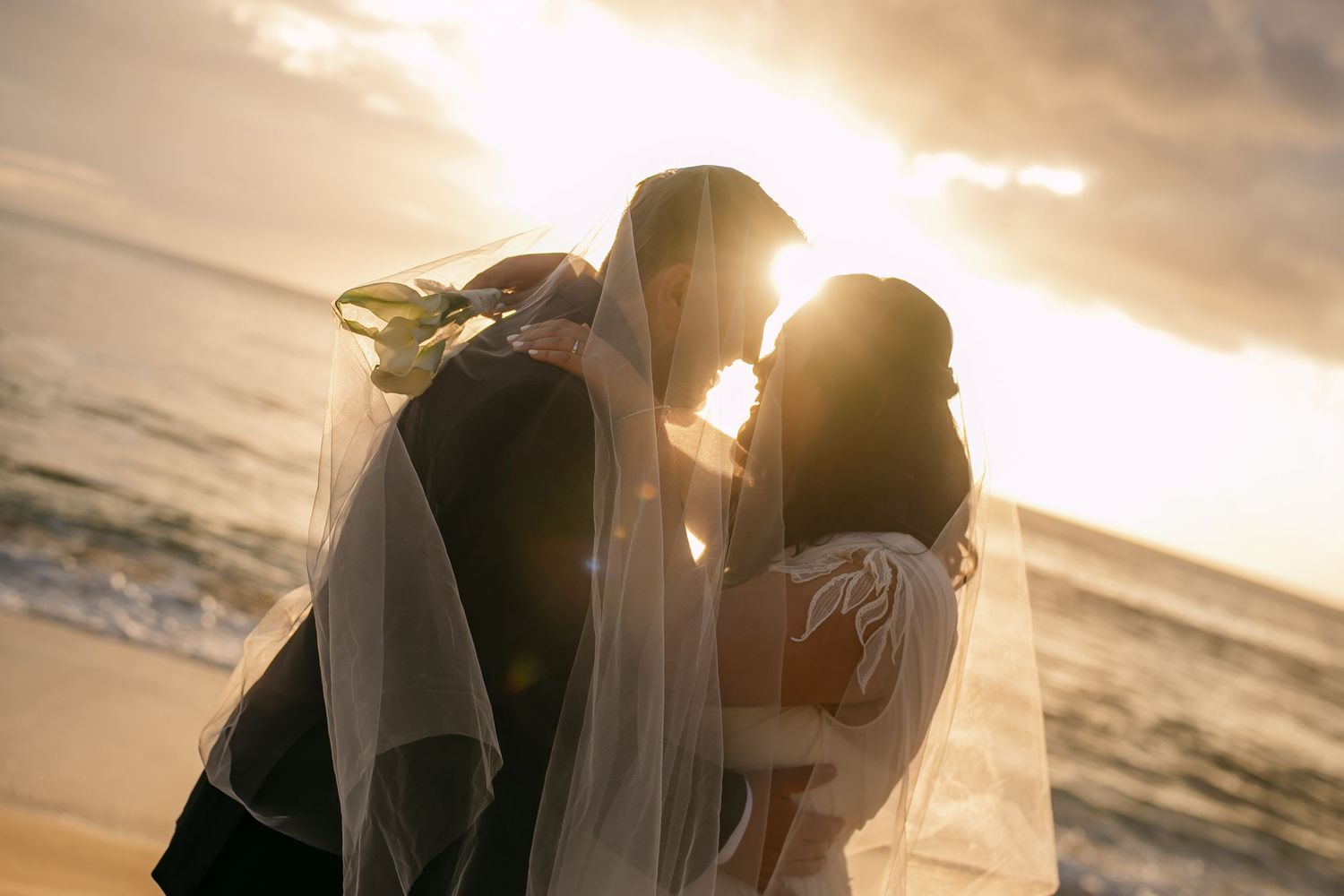 Bride and groom sharing a romantic kiss on the beach at sunset, with a flowing veil and golden light.