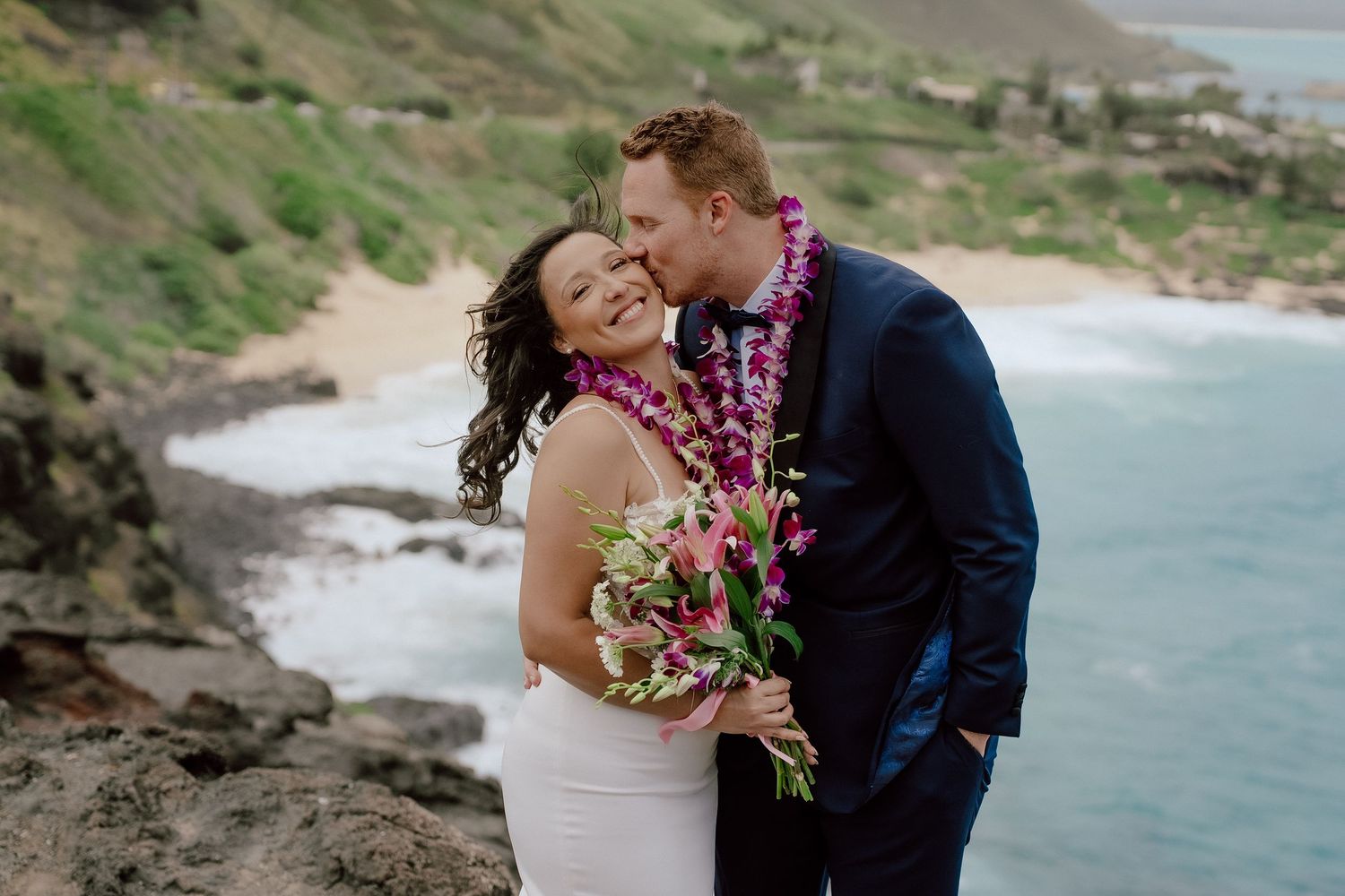 A couple in wedding attire share an intimate moment on a scenic Hawaiian cliffside overlooking the ocean.